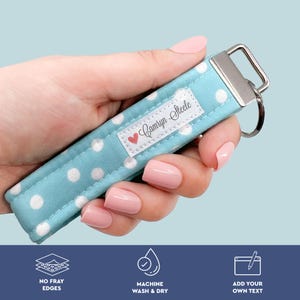 Peut inclure: Un porte-clés bleu clair à pois blancs. Le porte-clés a un anneau en métal argenté et une étiquette blanche avec le nom "Camryn Steele" et un cœur rouge. Le porte-clés est tenu dans une main.