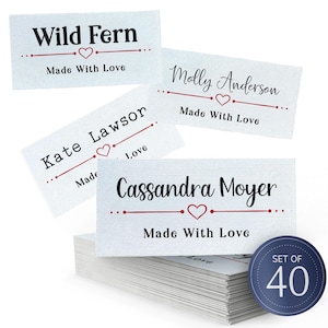 Può includere: Etichette in tessuto bianco con testo nero, cuori rossi e la scritta «Made With Love». Le etichette presentano nomi come «Wild Fern», «Molly Anderson», «Kate Lawson» e «Cassandra Moyer». È visibile una pila di etichette, con un cerchio blu che indica un set di 40.