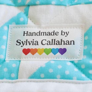 May include: A rectangular white fabric label stitched onto a light blue and white polka dot quilted textile. The label features the black text Handmade by Sylvia Callahan above a row of six small hearts in red, orange, yellow, green, blue, and purple.