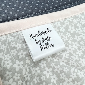 以下が含まれることがあります： 白い布製の衣類ラベルには黒い文字でHandmade by Kate Millerと印刷されており、グレーの花柄のキルティング生地と、白い水玉模様のダークグレーの生地の間に縫い付けられています。生地の間には淡いピンクのトリムがあしらわれています。