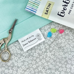 May include: A white fabric clothing label with the text Made with Love by Jamee Robinson and a rose illustration, placed on light green and white floral fabric next to gold stork scissors and three colorful bird-shaped sewing pins.
