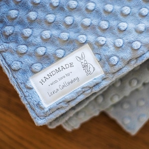 May include: A light blue minky blanket with a raised dot texture. A white rectangular fabric label is sewn onto the corner, featuring a black line drawing of a rabbit and the text: HANDMADE with love by Lena Calloway.