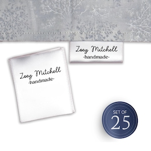 Può includere: Due etichette per abbigliamento in raso bianco con il testo stampato in nero Zoey Mitchell -handmade-, una attaccata a un tessuto grigio con un motivo a rami bianchi e una sciolta, accanto a un badge circolare blu scuro con il testo SET OF 25.