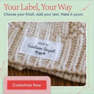 May include: A white rectangular fabric label sewn onto a beige knitted garment. The label features the black printed text "Made by Carolann August" with a decorative flourish underneath. The image includes the text "Your Label, Your Way" and "Customize Now".