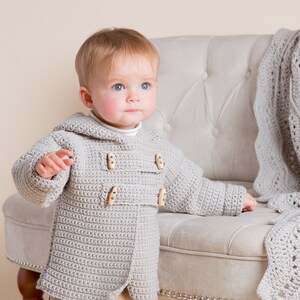 Puede incluir: Un bebé con una chaqueta de punto de ganchillo gris con botones de madera. El bebé está de pie frente a una silla blanca con una manta de ganchillo encima.