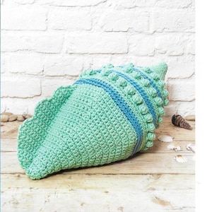 以下が含まれることがあります： ターコイズブルーとブルーの糸で編まれた貝殻の形をしたクッション。クッションは、テクスチャのある編み方で、立体的なデザインになっています。クッションは木製の表面の上に置かれています。