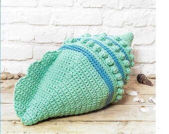 May include: A crocheted seashell pillow in shades of teal and blue. The pillow is made with a textured stitch and has a raised design. The pillow is sitting on a wooden surface.