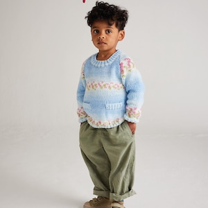 May include: A young child wearing a light blue and white knit sweater with pink floral accents and two front pockets. The child is also wearing olive green pants and tan shoes. The text "Hayfield" is at the top, and "Baby Blossom Chunky" is at the bottom.