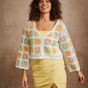 Könnte beinhalten: Eine weiße Häkeljacke mit einem Granny-Square-Muster in Gelb-, Rosa-, Blau- und Orangetönen. Die Jacke hat einen V-Ausschnitt und einen Knopfverschluss.