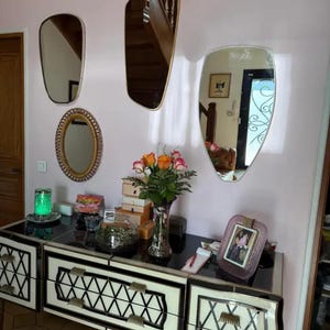 May include: A black and white dresser with decorative drawer fronts, topped with a variety of items including a vase of flowers, a terrarium, and a framed photo. Several mirrors of different shapes hang on the pink wall.