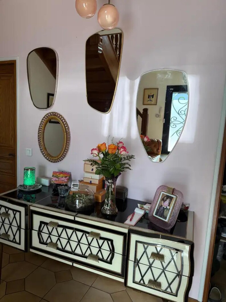 May include: A black and white dresser with decorative drawer fronts, topped with a variety of items including a vase of flowers, a terrarium, and a framed photo. Several mirrors of different shapes hang on the pink wall.