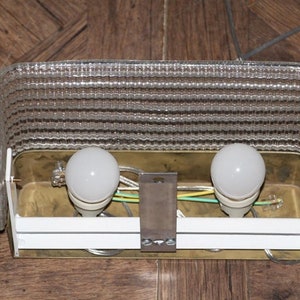 Puede incluir: Una l&aacute;mpara de lat&oacute;n con dos bombillas blancas y una pantalla de vidrio transparente con textura. La l&aacute;mpara tiene un respaldo de pl&aacute;stico blanco y est&aacute; cableada con cables verde, blanco y negro.