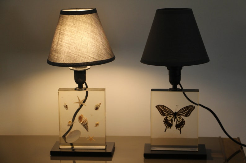 May include: Two table lamps with black lampshades. The lamp on the left has a clear glass base with seashells embedded in it. The lamp on the right has a clear glass base with a butterfly embedded in it.