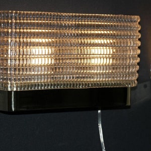 Puede incluir: Dos apliques de pared de lat&oacute;n con pantallas de vidrio transparente. Las pantallas tienen un dise&ntilde;o texturizado y acanalado y est&aacute;n iluminadas por bombillas.