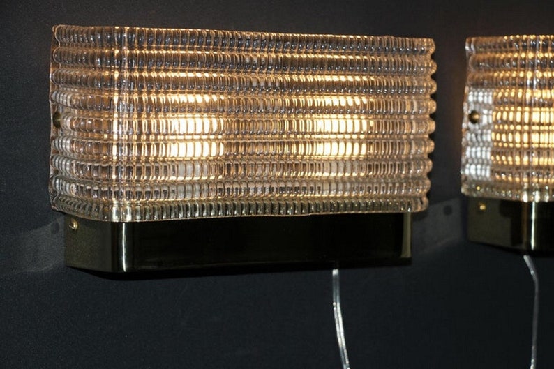 Puede incluir: Dos apliques de pared de lat&oacute;n con pantallas de vidrio transparente. Las pantallas tienen un dise&ntilde;o texturizado y acanalado y est&aacute;n iluminadas por bombillas.