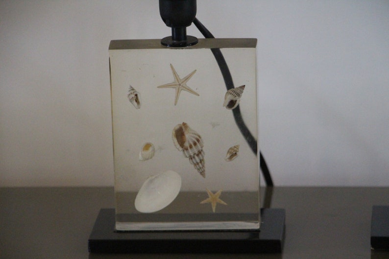 May include: A clear acrylic lamp base with embedded seashells and starfish. The lamp base is on a black base.