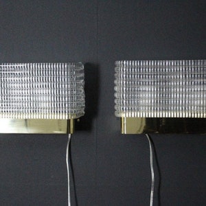 Puede incluir: Dos apliques de pared de lat&oacute;n y vidrio transparente con un patr&oacute;n de rejilla. Las l&aacute;mparas tienen un dise&ntilde;o sencillo y son perfectas para a&ntilde;adir un toque de estilo moderno a cualquier habitaci&oacute;n.