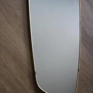 May include: A rectangular mirror with a soft, rounded shape and a thin, gold-colored frame. The mirror is mounted on a wooden surface with a herringbone pattern. The mirror's reflective surface is smooth and clear.