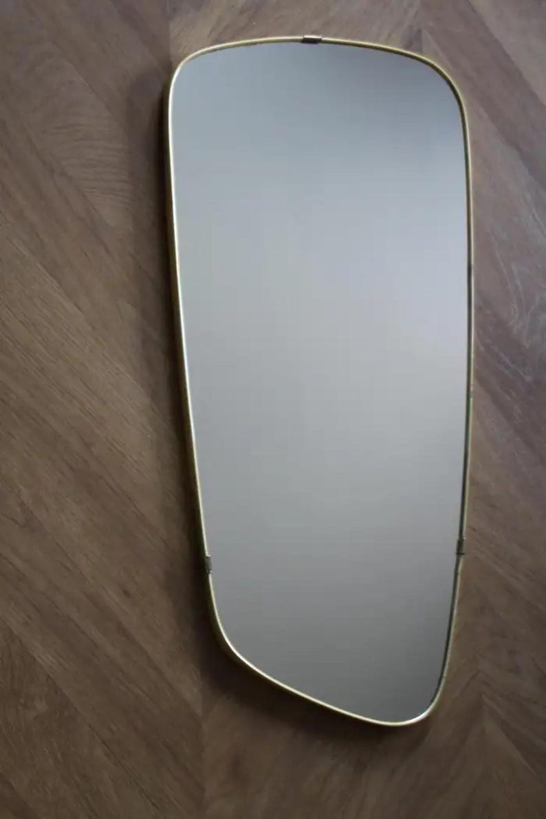 May include: A rectangular mirror with a soft, rounded shape and a thin, gold-colored frame. The mirror is mounted on a wooden surface with a herringbone pattern. The mirror's reflective surface is smooth and clear.