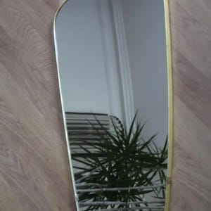 May include: A rectangular mirror with a soft, curved top and a gold-colored frame. The mirror reflects a room with a plant and a window. The mirror is hanging on a wooden wall.