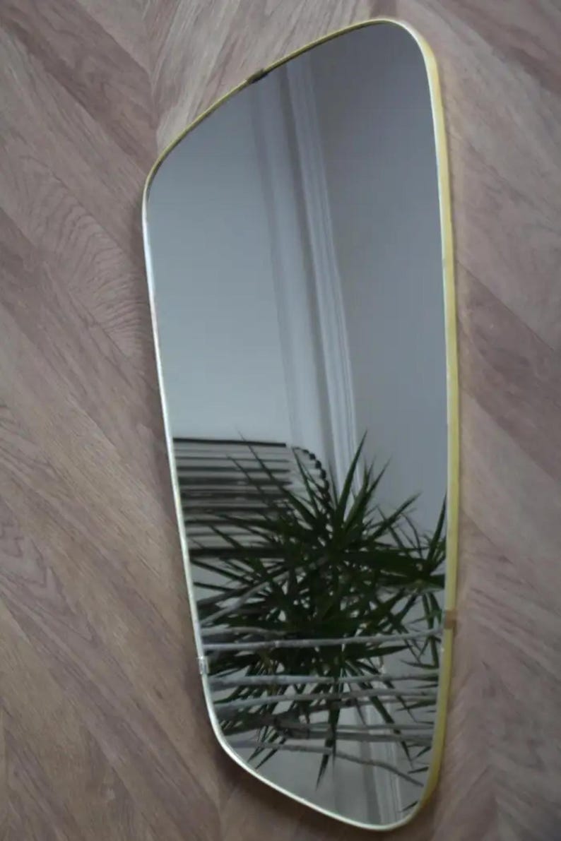 May include: A rectangular mirror with a soft, curved top and a gold-colored frame. The mirror reflects a room with a plant and a window. The mirror is hanging on a wooden wall.