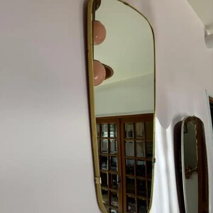 May include: A rectangular mirror with a gold-colored frame is mounted on a pink wall. The mirror has a unique, curved top edge. The reflection shows a room with wooden doors and a light fixture with two pink globes.