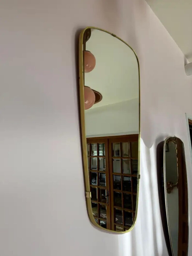 May include: A rectangular mirror with a gold-colored frame is mounted on a pink wall. The mirror has a unique, curved top edge. The reflection shows a room with wooden doors and a light fixture with two pink globes.