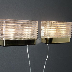 Puede incluir: Dos apliques de pared de lat&oacute;n con pantallas de vidrio transparente acanalado. Las luces est&aacute;n encendidas e iluminan la habitaci&oacute;n.