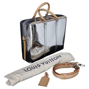 May include: A silver-colored travel bag with tan leather handles and trim. The bag has a zipper closure and embossed pattern. Includes a matching shoulder strap, dust bag with "LOUIS VUITTON" text, and a luggage tag.