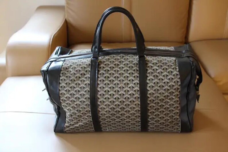 Puede incluir: Una bolsa de viaje negra y marr&oacute;n con un patr&oacute;n geom&eacute;trico. La bolsa tiene dos asas superiores y un cierre de cremallera.