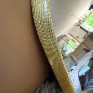 May include: Close-up of a gold-colored, textured, round mirror frame. The frame has a woven pattern and is set against a neutral-toned wall. The mirror reflects a glimpse of a room.