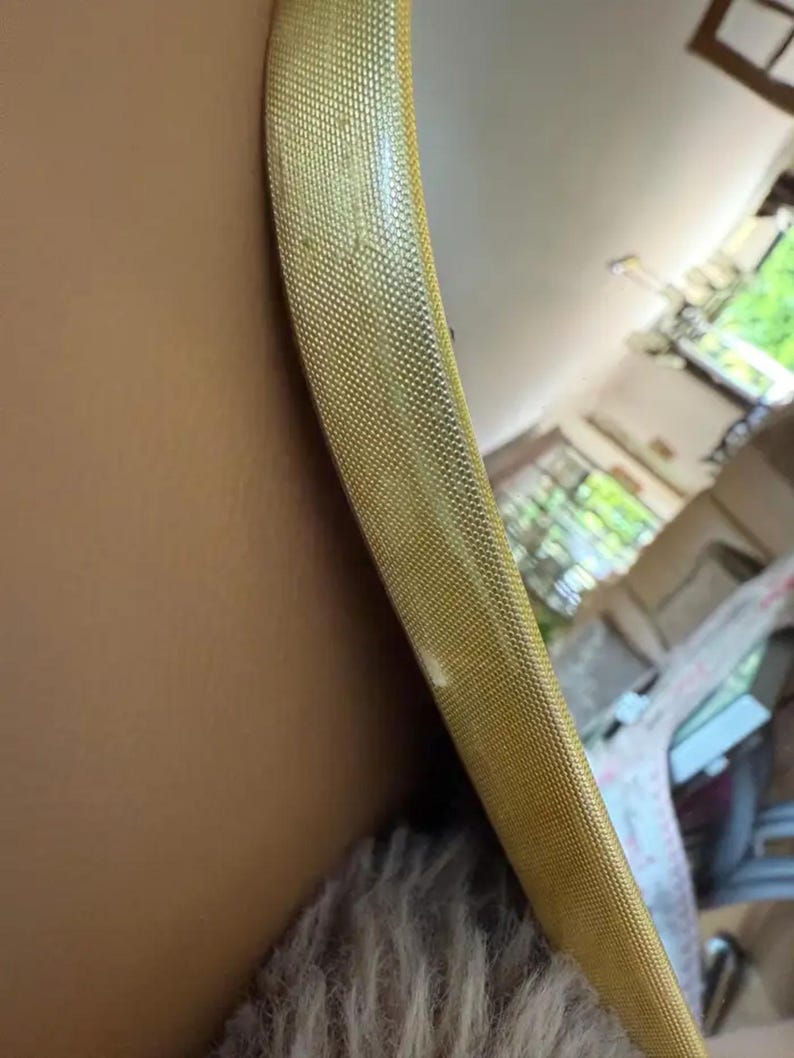 May include: Close-up of a gold-colored, textured, round mirror frame. The frame has a woven pattern and is set against a neutral-toned wall. The mirror reflects a glimpse of a room.