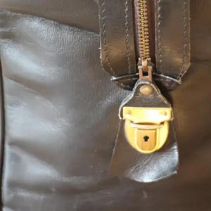 Puede incluir: Primer plano de una bolsa de viaje de cuero marr&oacute;n con una cremallera y un candado de lat&oacute;n. El candado tiene una cerradura.