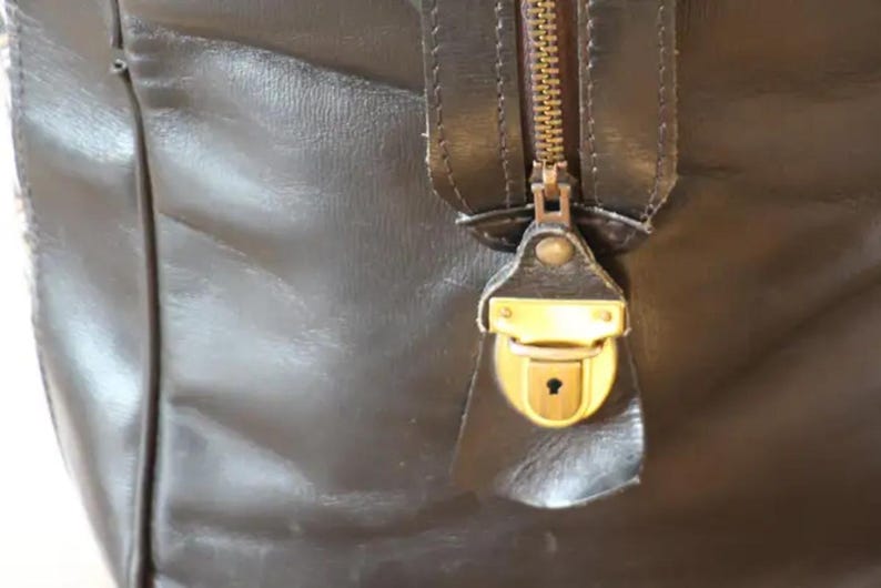 Puede incluir: Primer plano de una bolsa de viaje de cuero marr&oacute;n con una cremallera y un candado de lat&oacute;n. El candado tiene una cerradura.