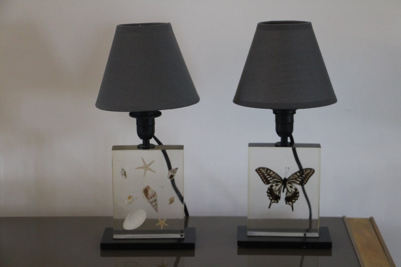 May include: Two table lamps with gray lampshades. The bases are clear acrylic blocks with a butterfly and seashells embedded inside.
