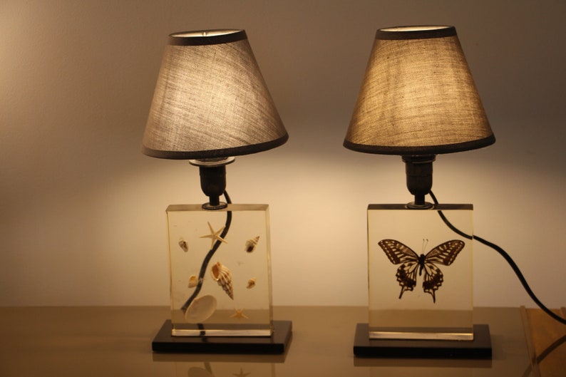 May include: Two table lamps with gray fabric lampshades. The bases are clear acrylic blocks with embedded decorative elements. One base features a butterfly, the other features seashells.