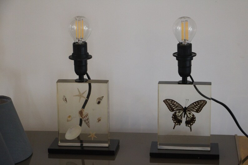May include: Two table lamps with clear glass bases. The base of the lamp on the left contains seashells and a starfish. The base of the lamp on the right contains a butterfly.