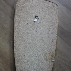 May include: The back of a rectangular, light brown wooden object with rounded corners. A small, white tag is attached near the top. The object appears to be a mirror or picture frame backing.