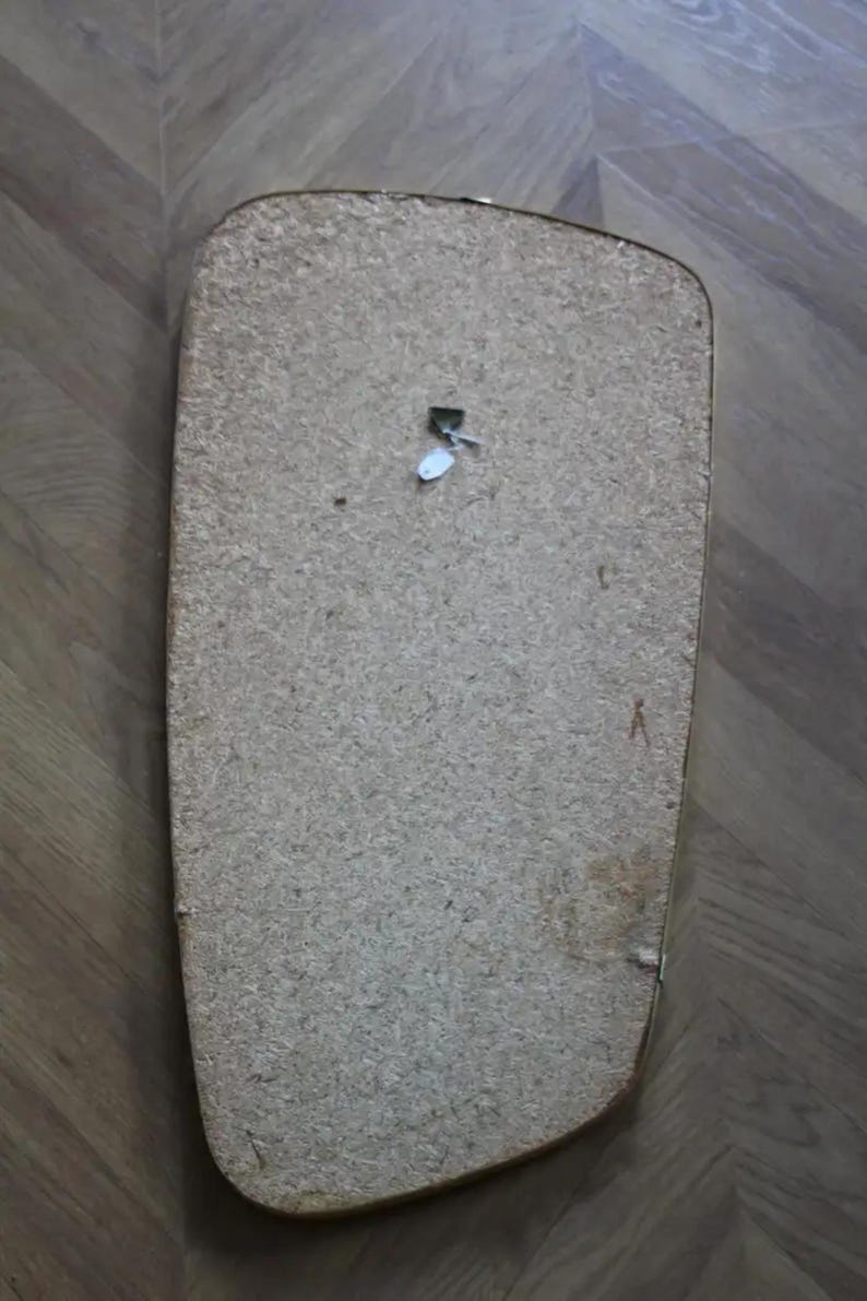 May include: The back of a rectangular, light brown wooden object with rounded corners. A small, white tag is attached near the top. The object appears to be a mirror or picture frame backing.