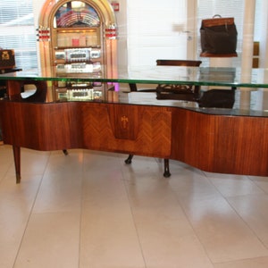 May include: A large, dark wood desk with a glass top. The desk has a curved front and sides, and the wood has a grain pattern. The desk is sitting on a light-colored floor.