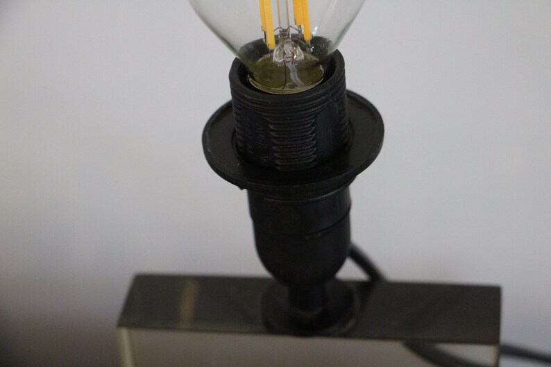 May include: Close-up of a black lamp socket with a clear light bulb inserted. The socket has a decorative ring around the base.