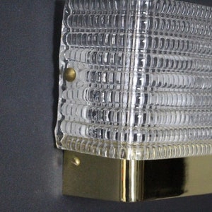 Puede incluir: Una l&aacute;mpara de pared de lat&oacute;n y vidrio transparente con una pantalla de vidrio texturizada. La l&aacute;mpara tiene dos tornillos en el lateral.