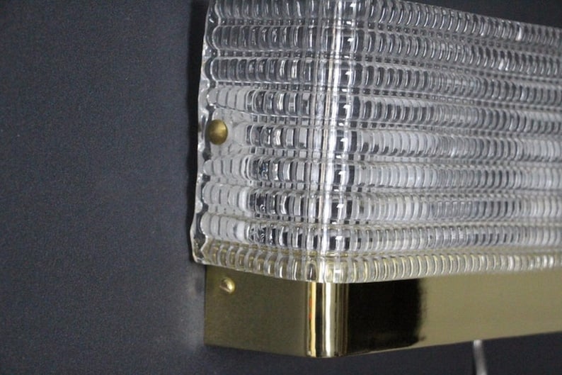 Puede incluir: Una l&aacute;mpara de pared de lat&oacute;n y vidrio transparente con una pantalla de vidrio texturizada. La l&aacute;mpara tiene dos tornillos en el lateral.