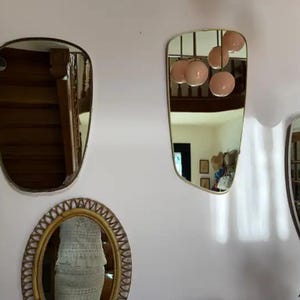 May include: A collection of decorative mirrors with gold frames and unique shapes hangs on a pale pink wall. One mirror is oval, another is a rounded rectangle, and a third has an asymmetrical shape. The mirrors reflect the interior of a room.