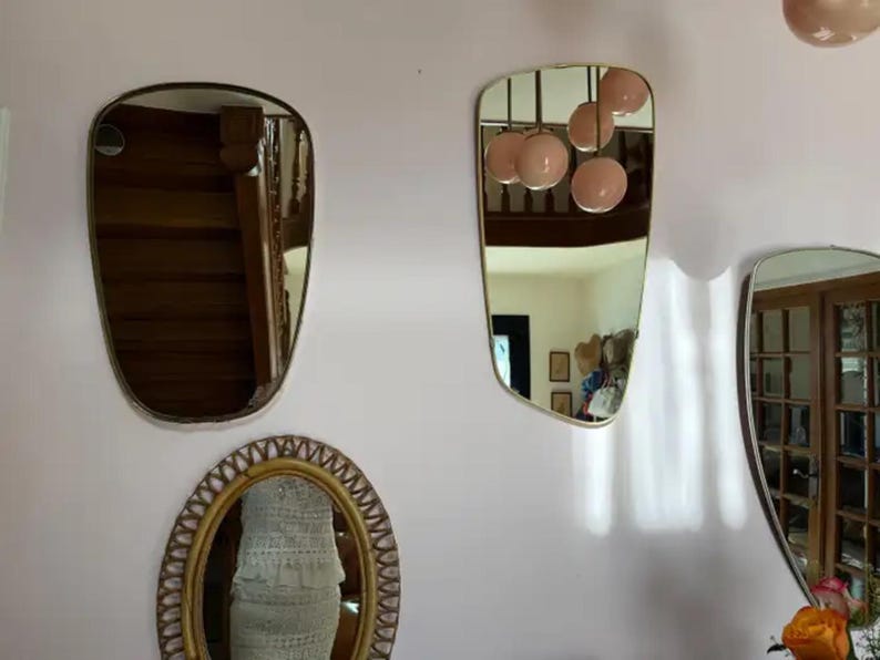 May include: A collection of decorative mirrors with gold frames and unique shapes hangs on a pale pink wall. One mirror is oval, another is a rounded rectangle, and a third has an asymmetrical shape. The mirrors reflect the interior of a room.