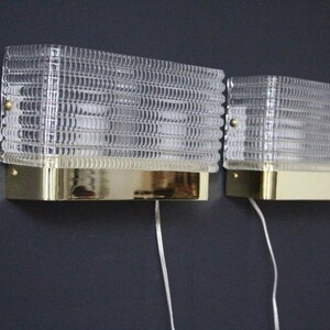 Puede incluir: Dos apliques de pared de lat&oacute;n con pantallas de vidrio transparente. Las pantallas tienen un patr&oacute;n geom&eacute;trico texturizado.