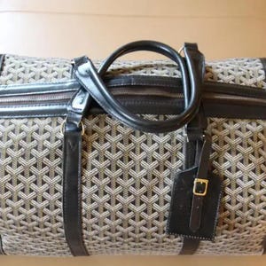 Puede incluir: Una bolsa de viaje de cuero negro y lona con un patr&oacute;n geom&eacute;trico. La bolsa tiene un asa superior y una etiqueta de equipaje desmontable.