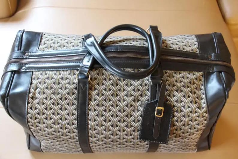 Puede incluir: Una bolsa de viaje de cuero negro y lona con un patr&oacute;n geom&eacute;trico. La bolsa tiene un asa superior y una etiqueta de equipaje desmontable.
