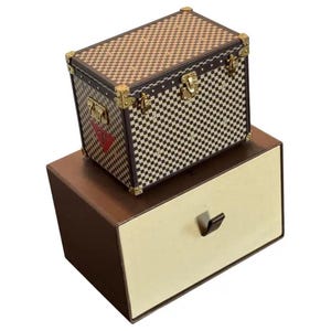 Puede incluir: Un baúl a cuadros marrón y crema con herrajes dorados y un emblema rojo, encima de una caja marrón y crema con un cajón. El baúl tiene forma rectangular y está diseñado con un patrón clásico.
