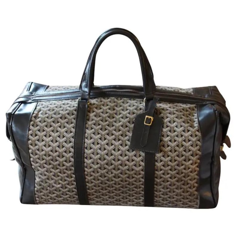 Puede incluir: Una bolsa de viaje negra y marr&oacute;n con un patr&oacute;n geom&eacute;trico. La bolsa tiene un asa superior y una etiqueta de equipaje.